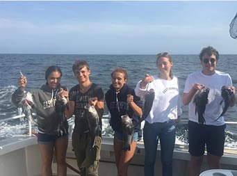 On a sunny day, 5 young adults smile and each hold up 2 sea bass.
