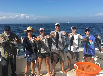 On a sunny day with a clear blue sky, a grandfather, two parents, and their triplet son and daughters all smile for the camera and hold out the sea bass they caught.
