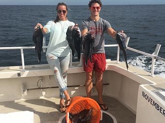 A man and woman smile and hold up 2 sea bass each, with more on the red mesh basket in front of them.