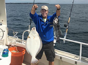 On a sunny day with the dark ocean behind him, a man wearing a royal blue fleece smiles and holds up a sea bass and jumbo fluke still on the line