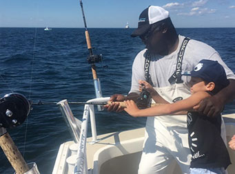 First mate, Gil, puts his arm around a young boy and helps him guide the use of a reel.