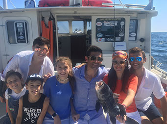 On a sunny day, 3 adults and 4 young adults sit together for a photo and hold up a large sea bass they caught.