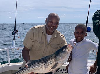 With the blue sky and ocean behind them, a holds up a striped bass standing next to a young boy and they both smile.