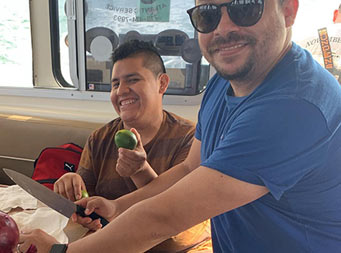 View inside the boat (in the galley) of two men preparing to make ceviche, with one man holding a whole lime, the other holding a whole red onion, and both smiling for the camera.