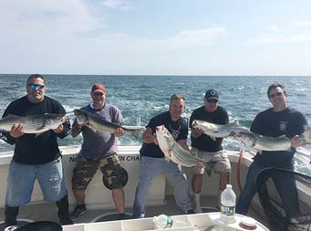 With the blue sky and ocean behind them, a group of 5 men each hold up striped bass.