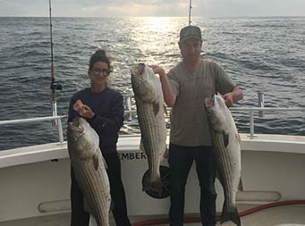 With the sun reflecting off the dark water behind them, a man holds up two striped bass and a woman holds up one.