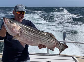 With the wake of the boat behind them, a man smiles and holds up the striped bass he caught.