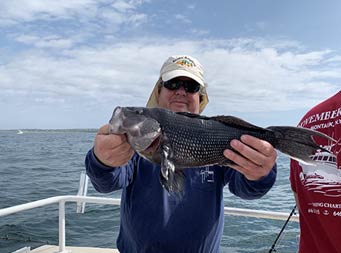 Under a Slightly cloudy sky, the camera takes a closeup of a man holding out the sea bass he caught.