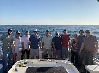 Ten men and women stand on the boat and pose for a photo with the ocean and sun behind them.