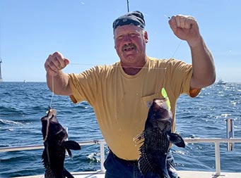 On a sunny day, a man wearing a bandana on his head and a yellow tshirt holds up a line with two sea bass on it.