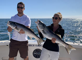 With a sunny clear blue sky above them, a man and woman, each wearing sunglasses, holds up the striped bass they caught .