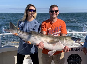 A man and woman both wearing sunglasses work together to hold up a striped bass using their arms and hands.