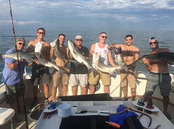 With clouds in the sky, but the sun shining upon them casting shadows late in the day, 7 men each hold up the striped bass they caught.