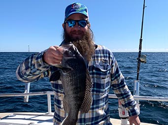 On a clear blue-sky day, a man smiles and holds up a sea bass.