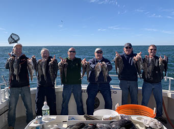 On a windy,sunny day with few clouds, 6 men wearing fall attired each hold up two sea bass.