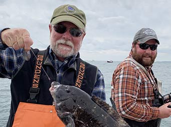 On a cloudy fall day, a man wearing sunglasses holds up the sea bass he caught while a man in the background fishing looks over.