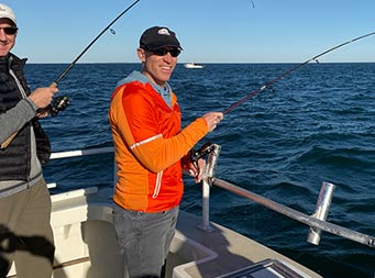 On a sunny, clear blue-sky day, a man wearing a blue hat, sunglasses and a red and orange windbreaker looks towards the camera as he holds onto his reel already in the water