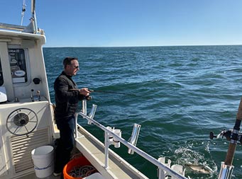 With the clear blue sky above and the water all around, the camera catches a man holding onto the reel, waiting for his catch off the side of the boat.