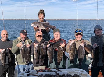 On a 
 sunny day with few clouds, Captain Jill stands in the background on the edge of the boat holding up one blackfish, while 6 men in front of her each hold up 2 sea bass, and more sea bass laid out on the filet table.