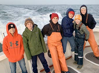 On a slightly overcast, chilly morning, 6 tween-age boys , some wearing hats and sweatshirts, pose together for a photo, with the wake of the boat behind them.