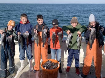 On a crisp, clear blue-sky day, 6 tween-age boys, some wearing hats, sweatshirts and fishing waders, each hold up 2 sea bass for the camera.