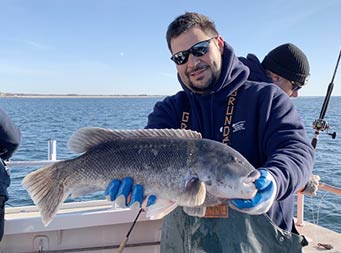 A man wearing sunglasses and a sweatshirt smiles and holds out a blackfish with two hands for the camera.
