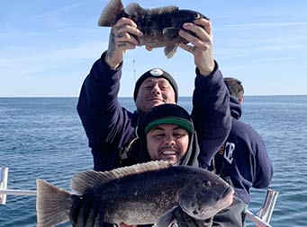 A man in the foreground smiles for the camera and holds up a large sea bass, while his friend stands directly behind him and holds up a much smaller sea bass over his head.