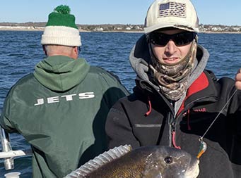 On a sunny and chilly morning, with the water and land in the background, a man bundled up and wearing sunglasses, holds up the blackfish (tautog) he caught. In the near background is a man facing the water wearing a green, NY Jets coat, which is facing the camera.