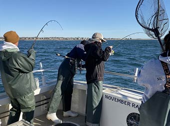 Three people, all wearing waders/bibs, hold onto their reels as their lines hang in the water. Off to the right is a glimpse of Captain Jill holding out a net ready to catch a fish.