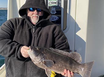 On a sunny, chilly morning, a man wearing a black sweatshirt and sunglasses, stands with his back against the boat and holds up a blackfish.