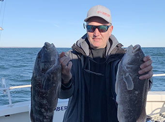 A man wearing a fleece, baseball cap and sunglasses, stands on the boat partly in the sun and partly in the shade, holding up a blackfish in each hand with more on the fillet table in front of him.