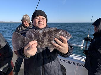 On a sunny, chilly morning, with the water behind him and land not too far in the distance, a man bundled up in a wool hat and winter coat smiles as he holds up the blackfish he caught.