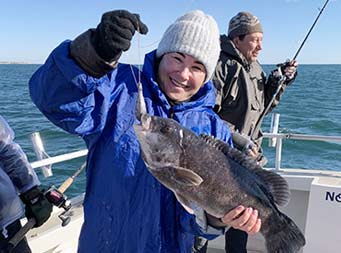 On a sunny, chilly morning, with the water behind them, a woman bundled up in a wool hat, gloves, winter coat and blue long raincoat over it, smiles big as she holds up a blackfish.