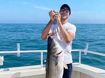 On a sunny day, a man wearing a hat and sunglasses holds up the striped bass he caught while a woman, wearing a green sweatshirt, stands behind him off to the right and smiles.