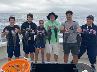 With the sky above filled with clouds and some light peeking through, two adults and 4 adolescent boys and girls stand together and each hold up 2 sea bass.