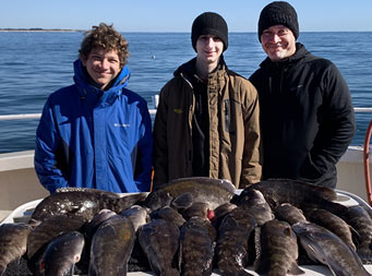 On a chilly, sunny day, a man and two young men wearing winter attire stand in front of the filet table where over a dozen blackfish are laid out