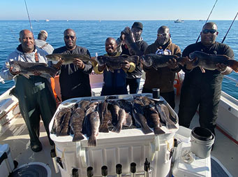 With a clear blue sky day and the blue ocean behind them, 6 men each hold up a blackfish, with more fish laid out on the filet table in front of them. First Mate Gil is in the background.