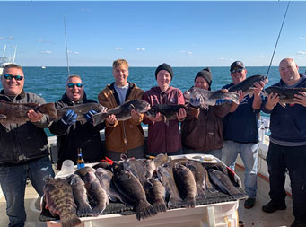 With a clear blue sky and few clouds, and the ocean behind them, 7 men and young men each hold up a blackfish, with more fish laid out on the filet table in front of them.
