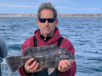 With the land and water behind him, a man wearing a maroon hoodie and sunglasses, holds up a blackfish.