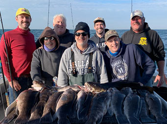 On a sunny, crisp day, seven men of varying ages wearing sweatshirts and hats, pose together with fish laid out on the filet table in front of them.