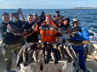 Eight men and women each hold fish up high and stand around the fillet table covered with fish.